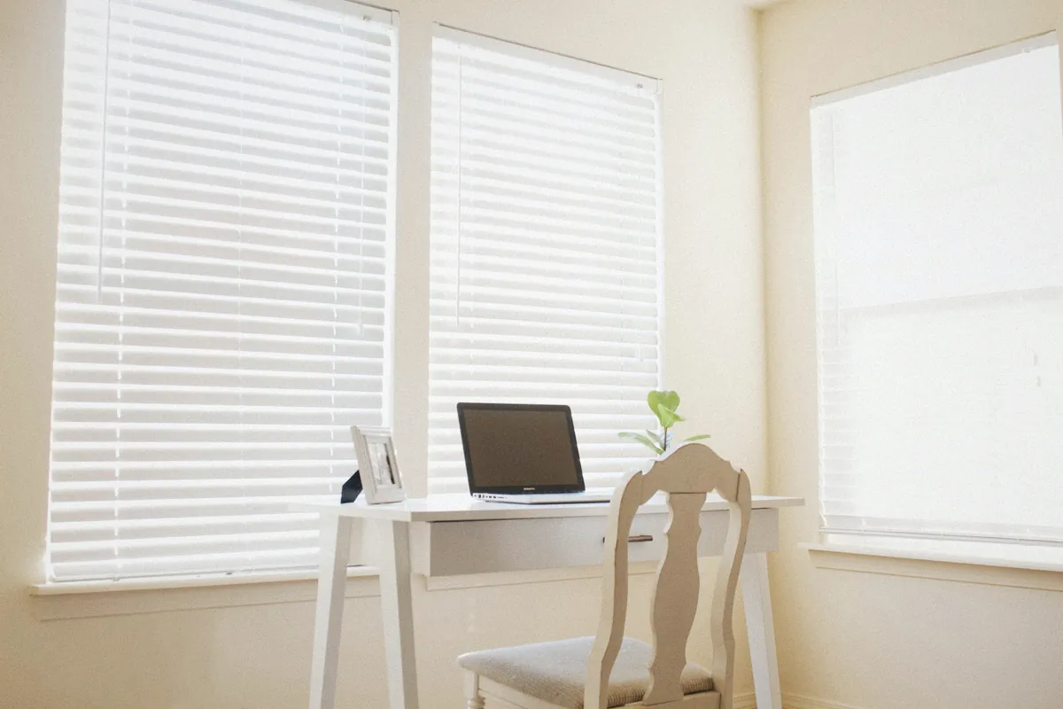 A laptop on a white desk in a bright home office with window blinds and a small plant